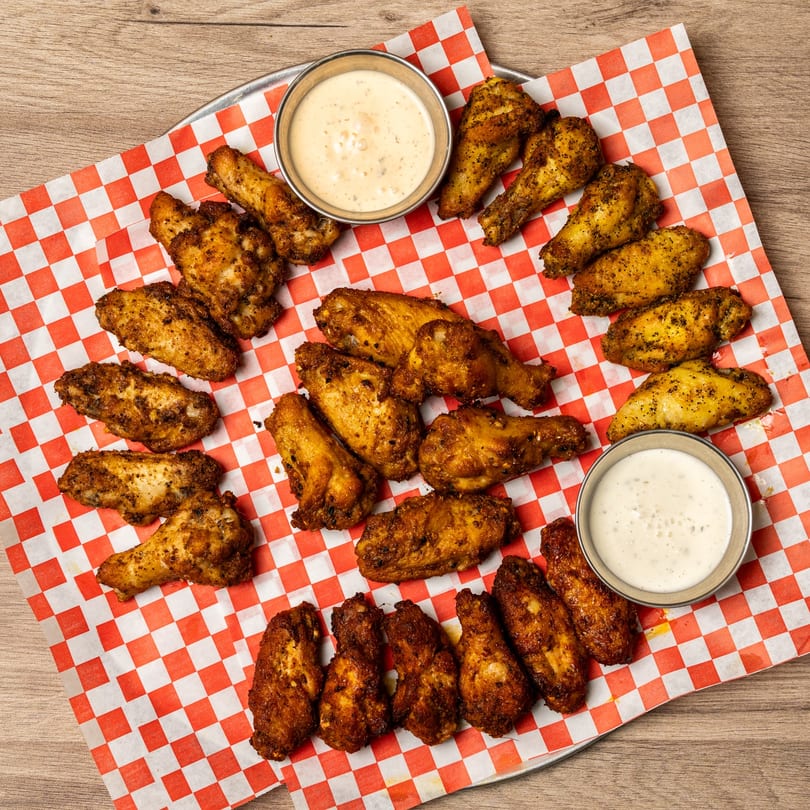 A platter of golden-brown chicken wings with two dipping sauces on red and white checkered paper.