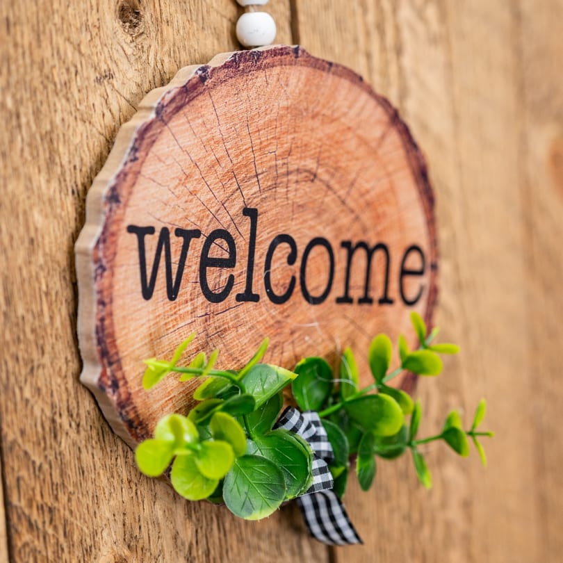 A wooden welcome sign with green leaves and a checkered bow hangs on a rustic wooden door.