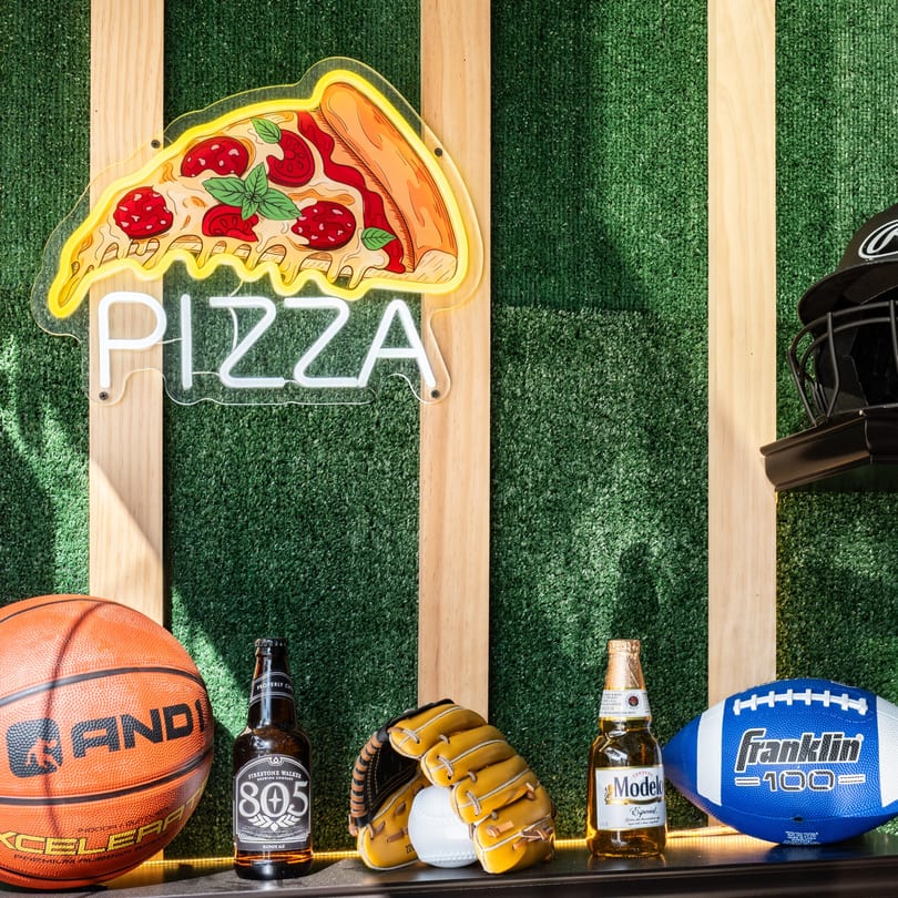 Neon pizza sign and sports equipment with beer bottles against a green turf wall.