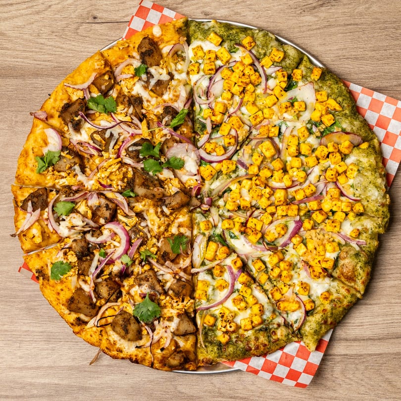 Half chicken tikka and half paneer tikka pizza with red onions and cilantro on a wooden table.
