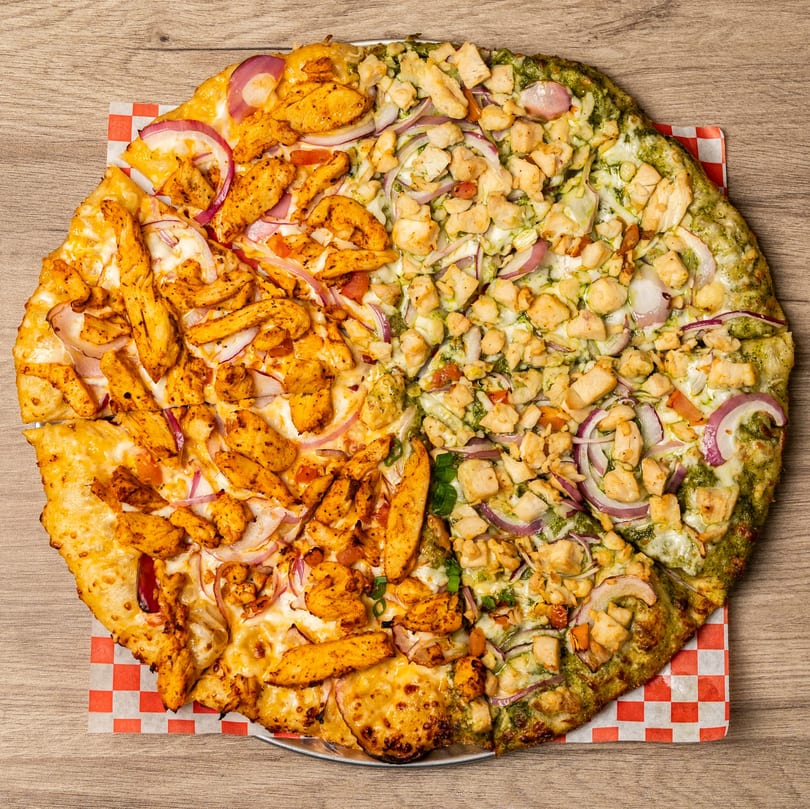 Half butter chicken and half pesto chicken pizza with red onions on a wooden table.